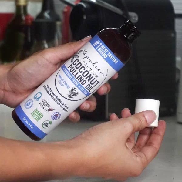 A person holding a bottle of Coconut Pulling Oil - Herbal Energy in their left hand and a white cap in their right hand. The bottle features various certifications and labels, including "#1 Best Tasting" and "Organic, Non-GMO, Vegan, Kosher and Cruelty-Free.