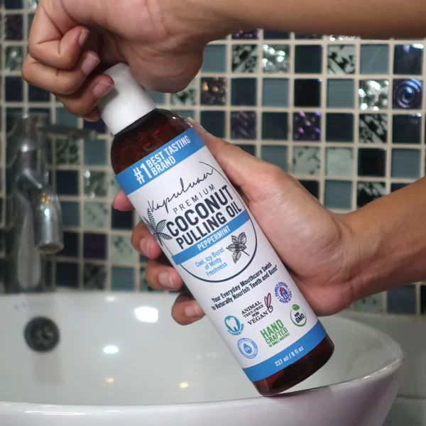 A person holds a bottle of "Coconut Pulling Oil - Peppermint" with their right hand in front of a tiled bathroom wall and sink. The bottle has a blue and white label and is marked as including peppermint and being hand-crafted, vegan, and non-GMO.