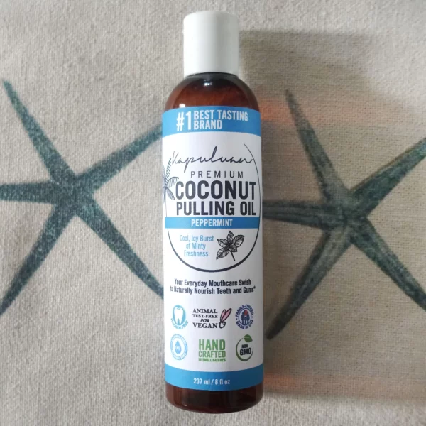 A brown bottle labeled "Coconut Pulling Oil - Peppermint" with peppermint flavor. The label promotes it as a premium product with fresh minty taste, vegan, cruelty-free, non-GMO, handcrafted, and environmentally friendly. It sits against a patterned background.