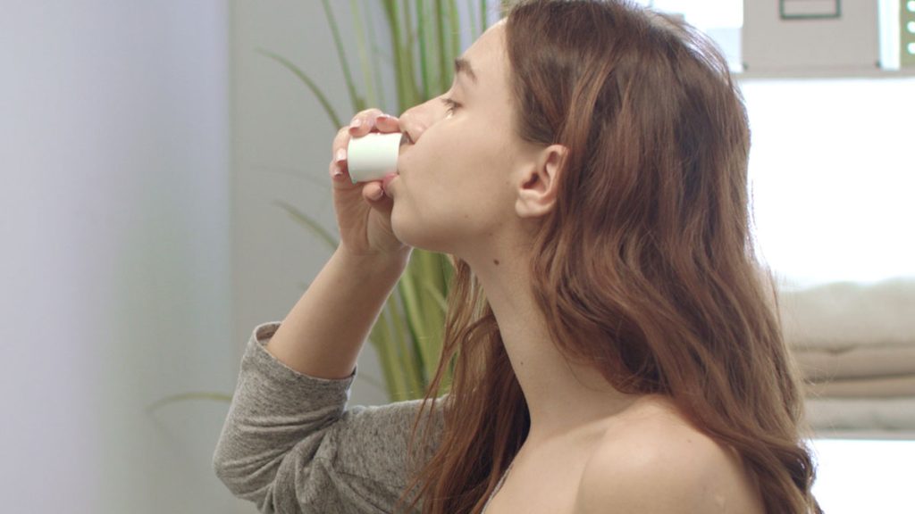 A young woman with long brown hair, wearing a grey top, is tilting her head back while drinking from a small white cup of Coconut Pulling Oil - Cinnamon. In the background, there is a blurred plant, towels, and some shelves.