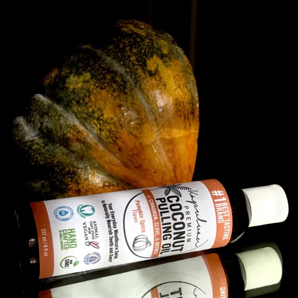 A bottle of coconut oil labeled "Coconut Pulling Oil - Pumpkin Spice" lies horizontally next to a green and orange gourd. The bottle features certifications such as USDA Organic, Vegan, and Non-GMO Project Verified.