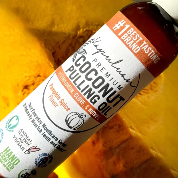 A bottle of Coconut Pulling Oil - Pumpkin Spice with a cinnamon, clove, and nutmeg flavor is placed against a bright orange background resembling a cut pumpkin. The label indicates it's pumpkin spice flavor and highlights features like vegan-friendly and handcrafted.