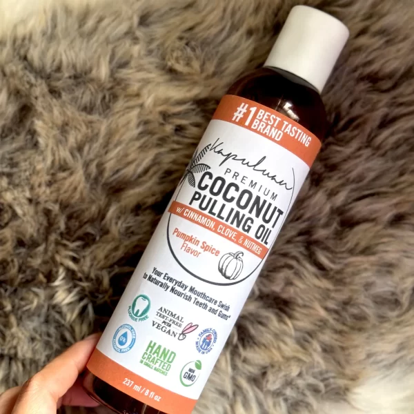 A bottle of Coconut Pulling Oil - Pumpkin Spice is held against a fur-textured background. The bottle showcases certifications like Vegan, Cruelty-Free, Non-GMO, and Handcrafted.