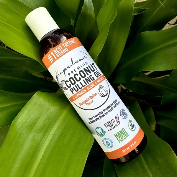A bottle of Coconut Pulling Oil - Pumpkin Spice rests on a green leaf. The bottle features several badges, including vegan, non-GMO, and cruelty-free. It is labeled "#1 Best Tasting Brand" and "Handcrafted in the USA." The bottle size is 237 ml.