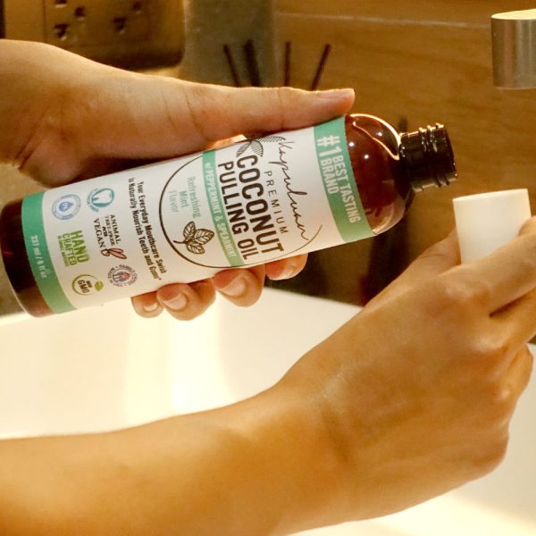 A person is pouring coconut pulling oil from a brown bottle labeled "Coconut Pulling Oil - Refreshing Mint" into a plastic cap. The person is standing by a sink. Various certifications and claims are visible on the bottle label.