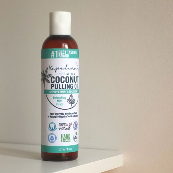 A bottle of Coconut Pulling Oil - Refreshing Mint stands on a white surface. The label highlights its peppermint and spearmint mix, vegan, cruelty-free, and handcrafted attributes, with logos indicating no GMO, gluten, or preservatives.