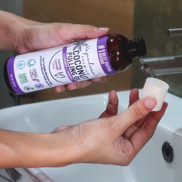 A pair of hands holding a brown bottle with a purple label reading "Coconut Pulling Oil - Soothing Lavender" while pouring some into a small white cap over a sink. The background appears to be a bathroom setting with a faucet and soap dish visible.