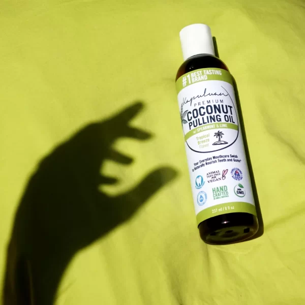 A bottle of Coconut Pulling Oil - Tropical Breeze rests on a bright yellow surface. There’s a large hand shadow reaching towards the bottle. The bottle label displays various certifications, including non-GMO, vegan, and cruelty-free.