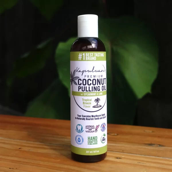 A bottle of Coconut Pulling Oil - Tropical Breeze sits on a wooden surface with green leaves in the background. The label highlights it as a "Tropical Breeze Flavor" and notes that it is natural, vegan, animal-friendly, and handcrafted.