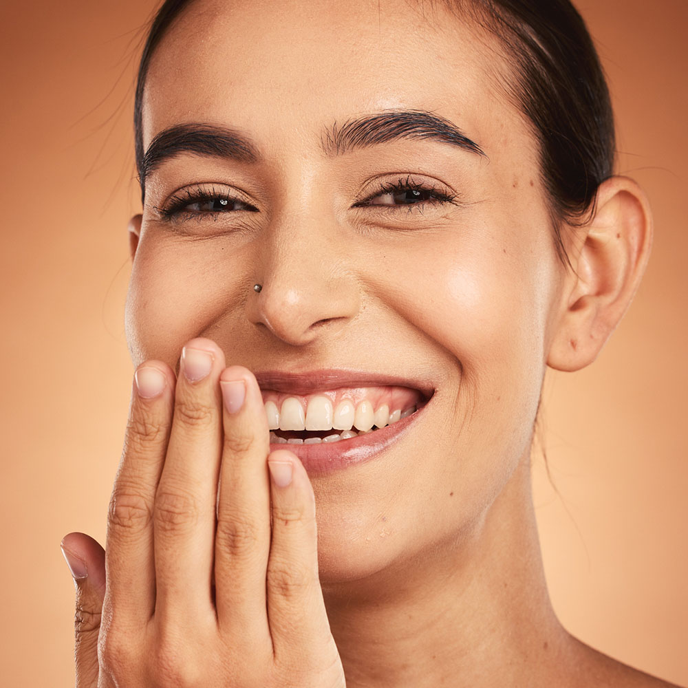 A person with smooth skin smiles warmly, covering their mouth with one hand, showing neatly manicured nails. The background is a soft, gradient shade of brown that complements their skin tone. Their hair is pulled back, emphasizing their face.