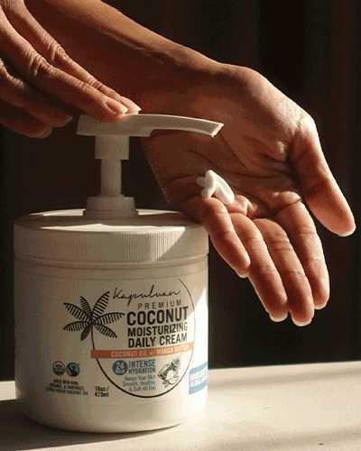 A hand is pressing a white pump dispenser, releasing a dab of cream infused with coconut oil into the palm of another hand. The product is labeled "Kapalua Premium Coconut Moisturizing Daily Cream," and the container features a black palm tree and product details.