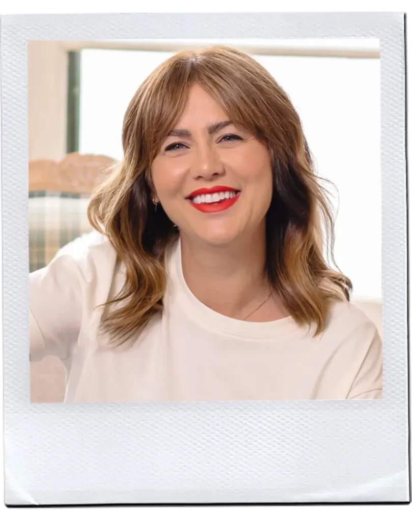 A woman with light brown, wavy hair and bangs, smiles widely showing her white teeth. She is wearing a white T-shirt and has bright red lipstick, her hair radiating a healthy shine that suggests she uses coconut oil. The photo has a Polaroid-style border. The background is softly blurred, suggesting an indoor setting with natural light.