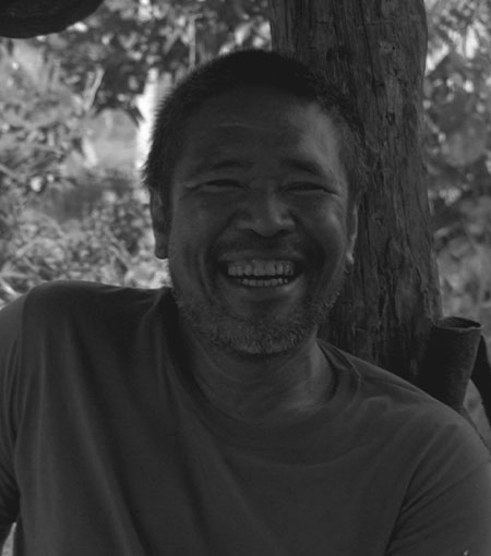 A grayscale photograph of a man with short hair and a beard smiling broadly. He wears a plain t-shirt and is leaning against a tree or wooden post. The background consists of blurred foliage, suggesting an outdoor setting.