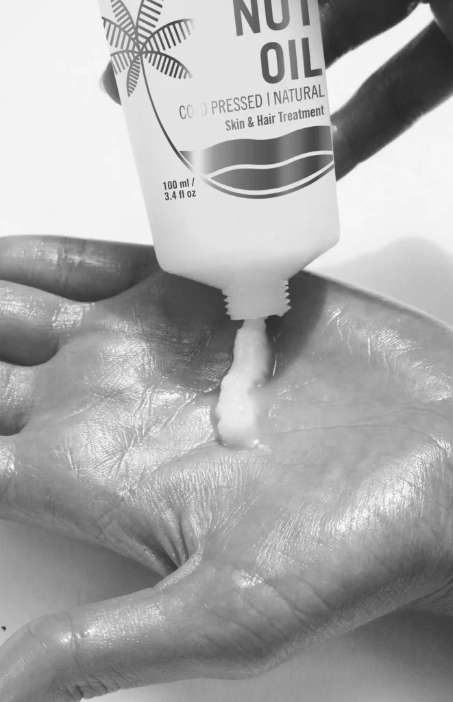 A person's hand is cupped to catch a creamy product being squeezed out of a bottle labeled "Nut Oil" with "Cold Pressed | Natural" and "Skin & Hair Treatment". The bottle is held by another hand, and the image is in black and white.