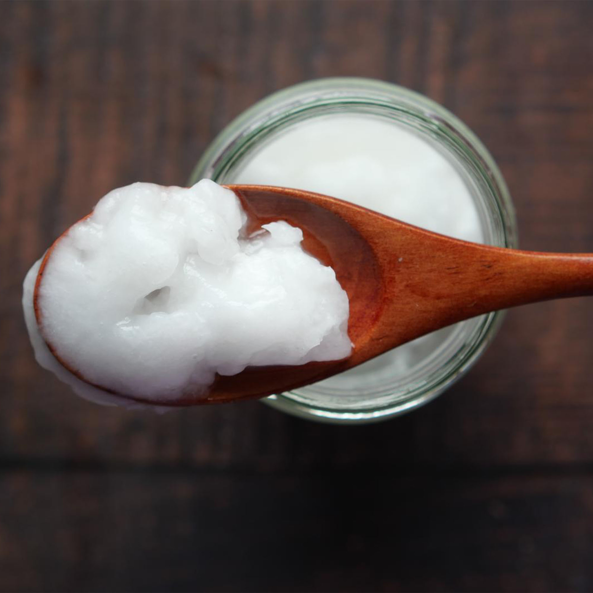 A wooden spoon holding a spoonful of white, creamy coconut oil is positioned over an open jar filled with more coconut oil. The background reveals a dark wooden surface.