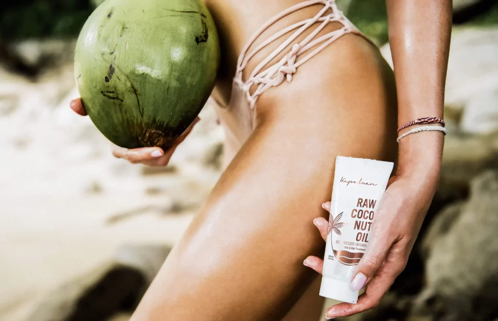 A person wearing a strappy bikini bottom is holding a green coconut in one hand and a tube labeled "Raw Coconut Oil" in the other. The scene features a tropical beach background with sand and rocks.
