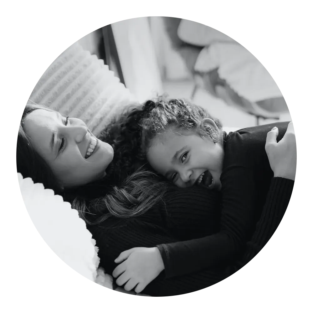 A woman and a young girl, both wearing dark clothing, share a joyful hug and laugh together. They are reclining comfortably on a cushioned surface. The image is in black and white and cropped into a circular shape.