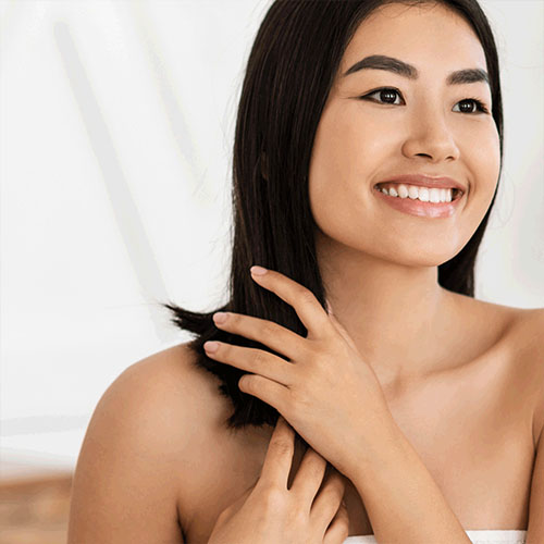 A smiling woman with straight black hair touches her shoulder. She has bright eyes and is looking off to the side. The background is softly blurred, creating a light and airy atmosphere.