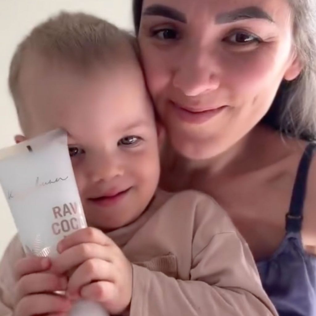 A woman and a child smiling at the camera. The child is holding a white tube labeled "RAW COCO" close to their face. They are both wearing light-colored clothing.