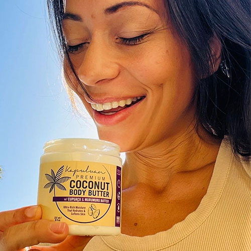 A person smiling and holding a jar of Kapuluan Premium Coconut Body Butter. The label highlights its moisturizing benefits with cupuaçu and murumuru butter. The background has a clear blue sky and palm leaves.