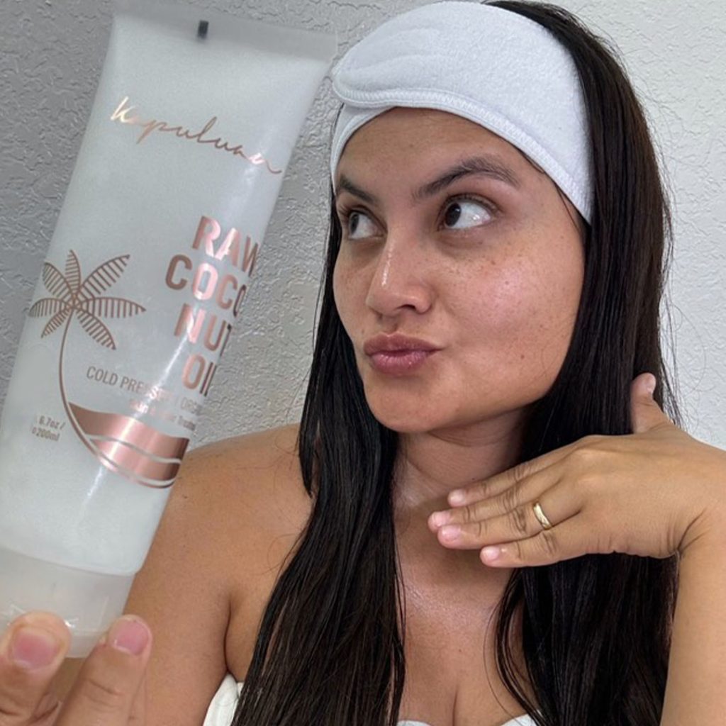 A person wearing a white headband holds a tube of raw coconut oil. They have long dark hair and are making a playful expression with their hand under their chin. The tube features a palm tree design.