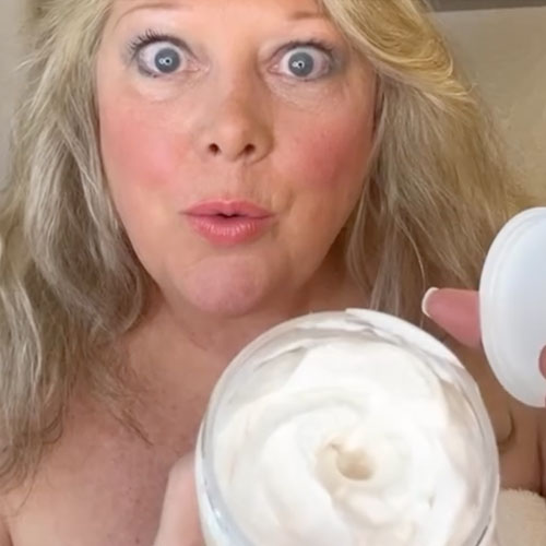 A woman with long blonde hair holds an open jar of cream close to the camera, looking surprised. She has makeup on and is indoors, with a neutral background.