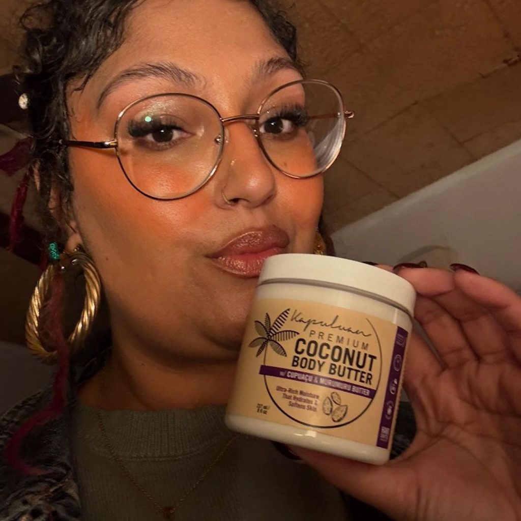 A person with glasses and large hoop earrings is holding a jar of coconut body butter. They are wearing a sweater with a patterned design and appear to be inside a room with a wooden ceiling.